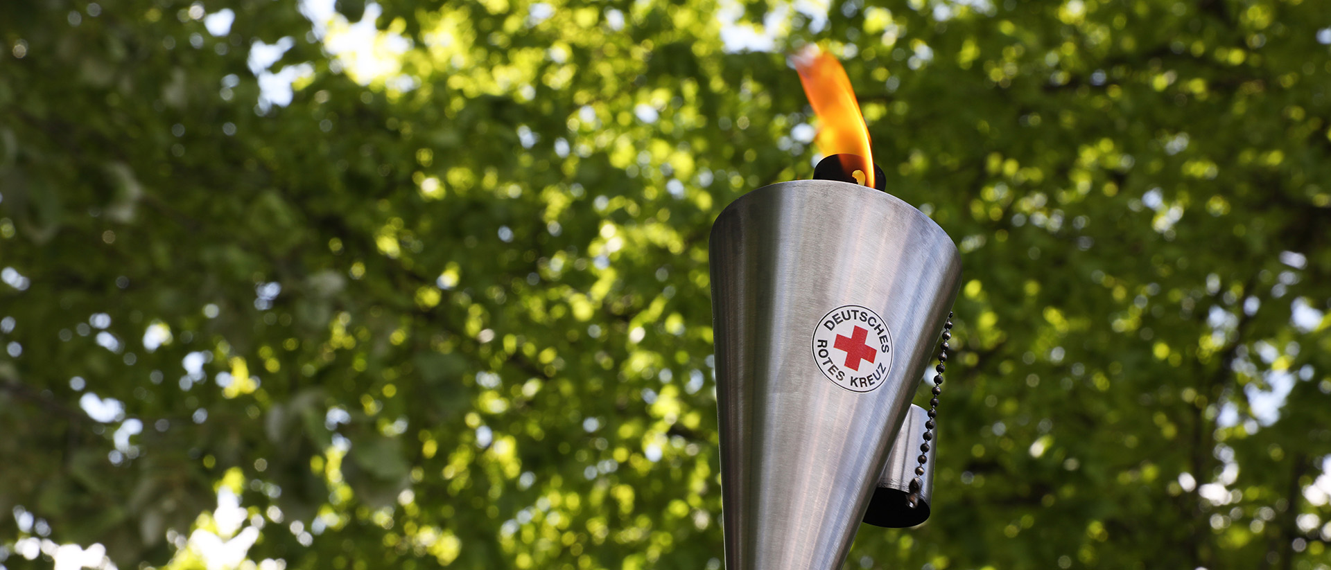 Eine brennende Fackel aus Edelstahl mit dem Logo des Deutschen Roten Kreuzes. Im Hintergrund sind grüne Baumkronen zu sehen.