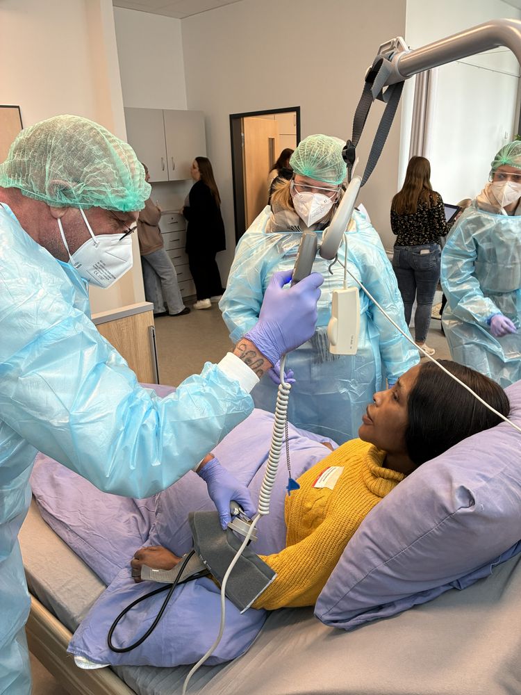 Um eine Person in einem Krankenbett stehen 3 Pflegekräfte in Schutzanzügen herum.