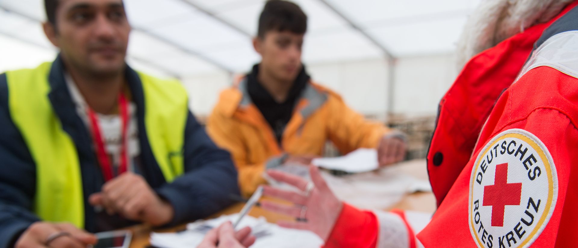 In einem großen Zelt spricht eine Person vom Deutschen Roten Kreuz mit zwei Menschen mit Migrationshintergrund.