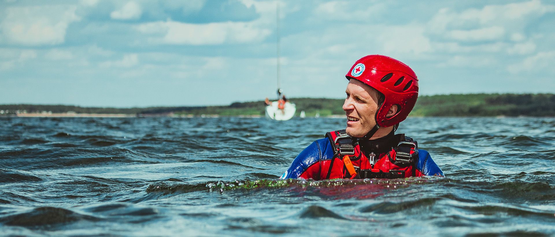 Ein junger Mann im Neopren-Anzug und rotem Helm schwimmt in einem See.