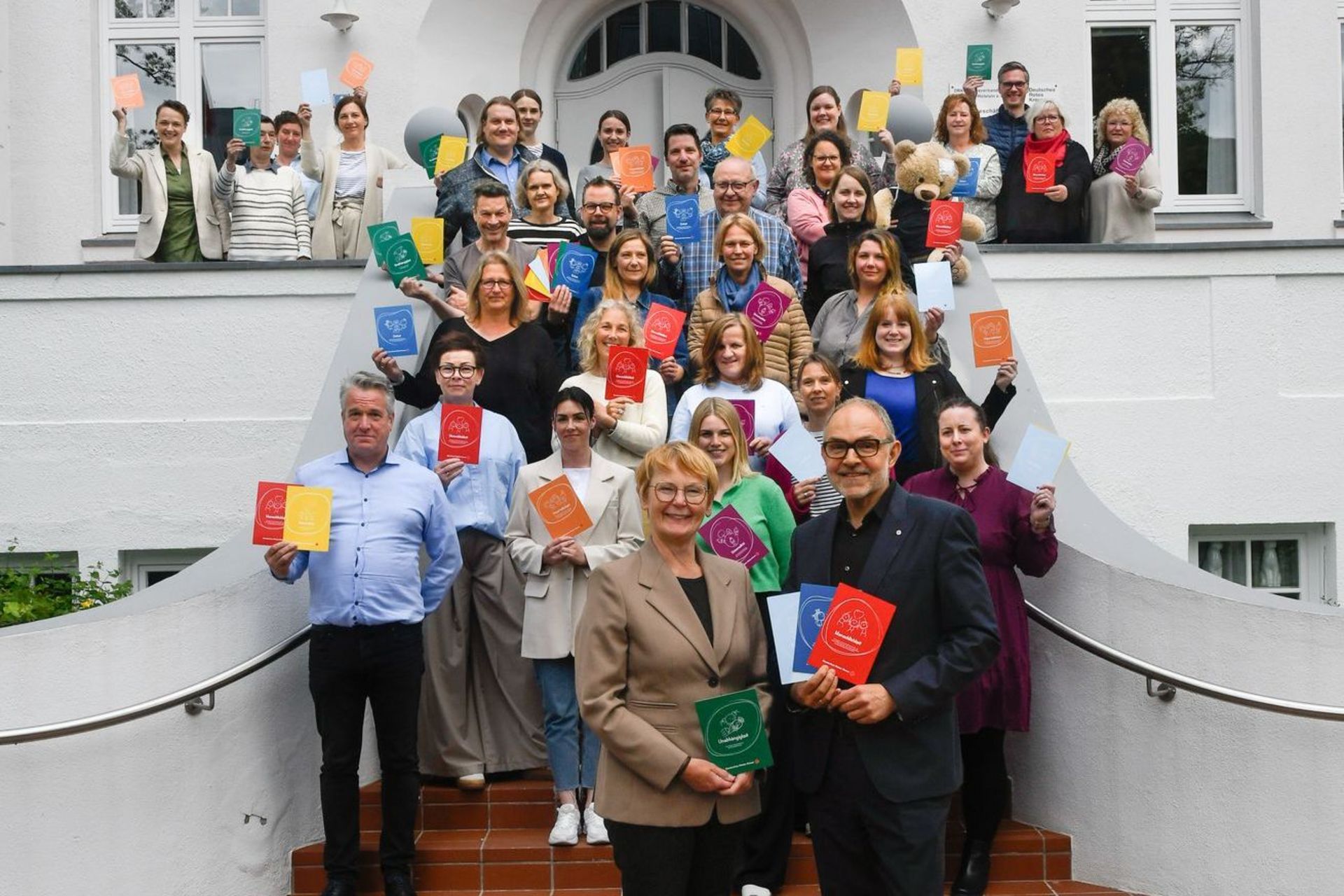 Gruppenfoto: Rund 40 Menschen stehen auf einer Treppe und halten bunte Karten in die Luft.