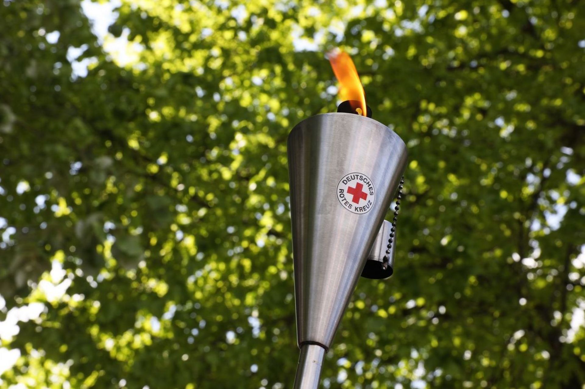 Eine brennende Fackel aus Edelstahl mit dem Logo des Deutschen Roten Kreuzes. Im Hintergrund sind grüne Baumkronen zu sehen.