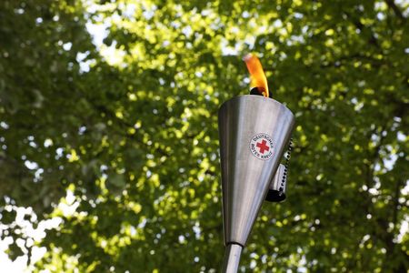 Eine brennende Fackel aus Edelstahl mit dem Logo des Deutschen Roten Kreuzes. Im Hintergrund sind grüne Baumkronen zu sehen.