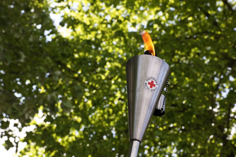 Fackellauf nach Solferino Eine brennende Fackel aus Edelstahl mit dem Logo des Deutschen Roten Kreuzes. Im Hintergrund sind grüne Baumkronen zu sehen.