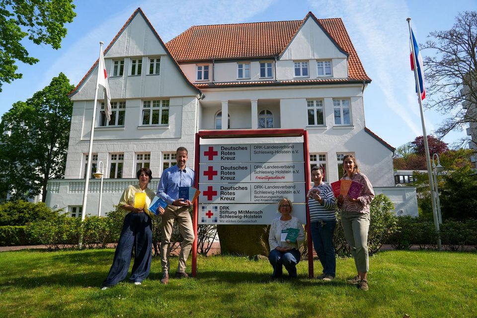 5 Personen stehen vor einem großen DRK-Schild. Im Hintergrund steht eine große weiße Altbauvilla: die Landesgeschäftsstelle des DRK Schleswig-Holstein.