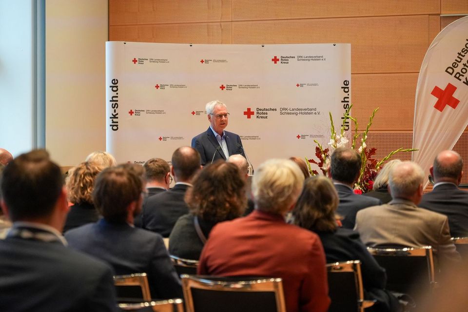 DRK-Präsident Torsten Geerdts Ein Mann im Anzug steht an einem Rednerpult und spricht zu einigen Menschen.