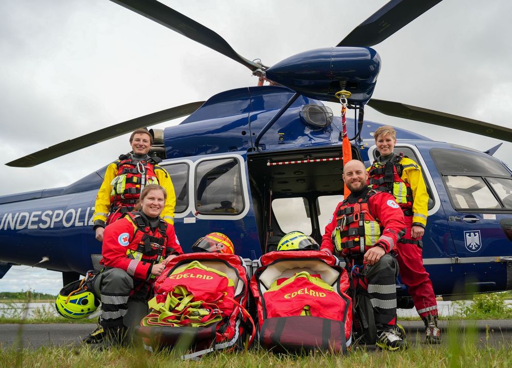 Vor einem blauen Bundespolizei-Hubschrauber stehen je zwei Einsatzkräfte von DRK und DLRG. Zwischen ihnen ist spezielles Rettungsmaterial platziert, darunter Rettungsdreiecke oder Baby-Rescue-Bags.
