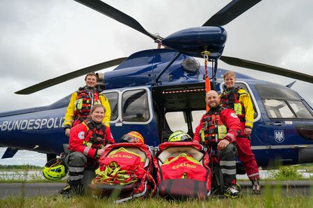 Vor einem blauen Bundespolizei-Hubschrauber stehen je zwei Einsatzkräfte von DRK und DLRG. Zwischen ihnen ist spezielles Rettungsmaterial platziert, darunter Rettungsdreiecke oder Baby-Rescue-Bags.