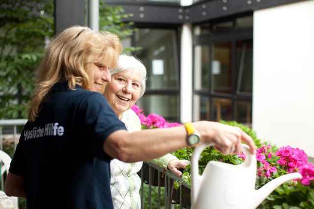 Eine lächelnde Seniorin steht auf ihrem Balkon neben einer DRK-Pflegekraft, die gerade rosafarbene Geranien gießt.