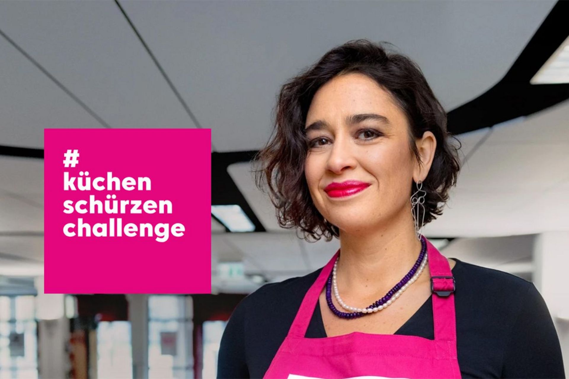 Eine junge Frau steht auf einem großen Büroflur. Sie trägt eine pinkfarbene Schürze. Neben ihr steht in einem pinkfarbenen Kasten: #küchenschürzenchallenge.