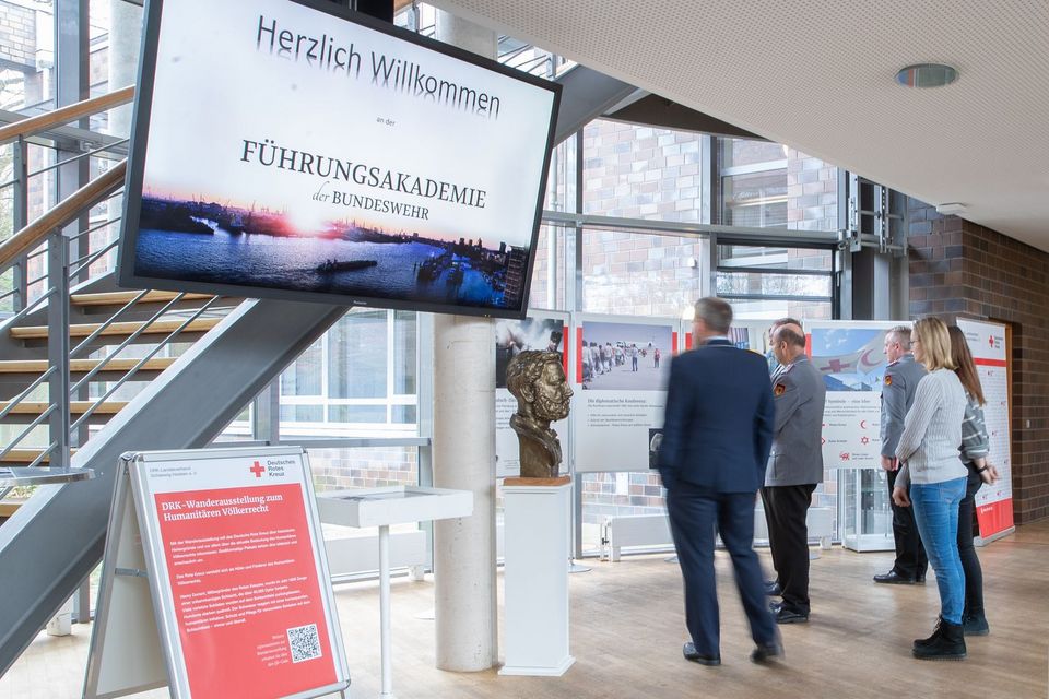 Sechs Personen stehen in einem Foyer und schauen sich dort eine Ausstellung an. Auf einem Plakat steht: DRK-Wanderausstellung zum Humanitären Völkerrecht.