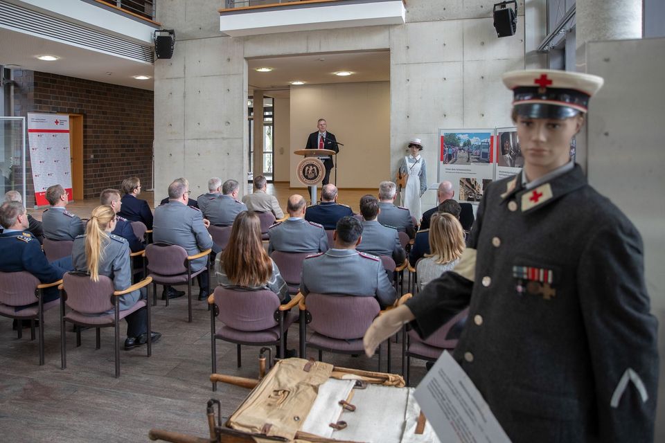 Eine Person steht an einem Rednerpult und spricht vor rund 20 Leuten. Im Vordergrund steht eine Puppe in Uniform und Rotkreuz-Mütze.