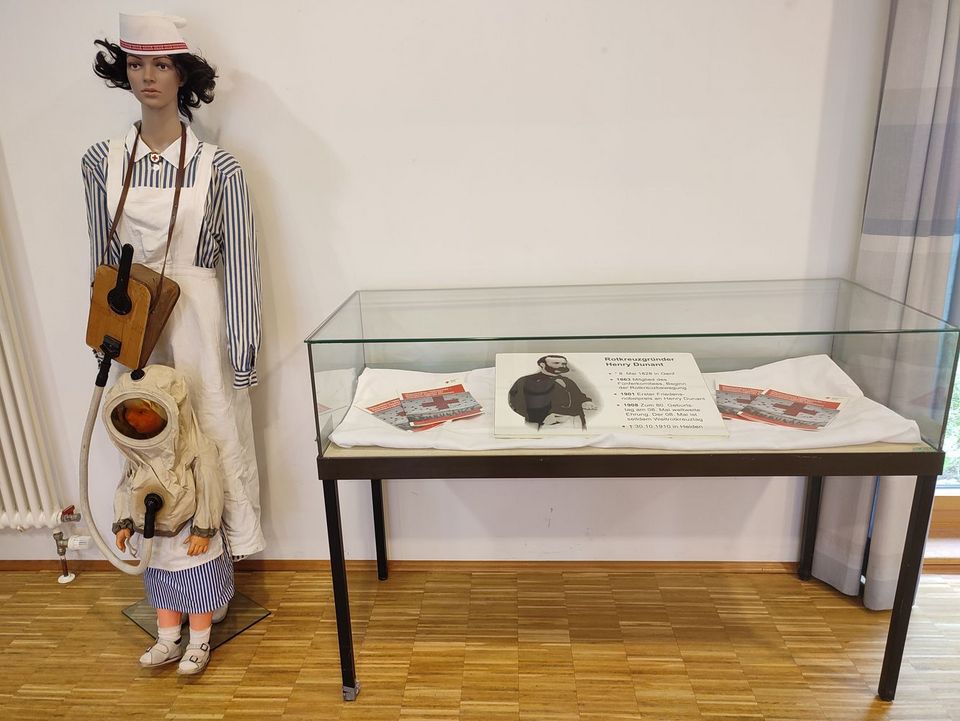 In einer Vitrine liegt eine Tafel mit Infos zu Henry Dunant. Links daneben steht eine Puppe in Rotkreuzschwestern-Kluft.