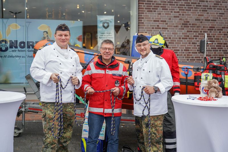 Drei Männer stehen vor einem Rettungsboot. Zwei von ihnen tragen eine weiße Koch-Jacke, der dritte eine rote Einsatzjacke.