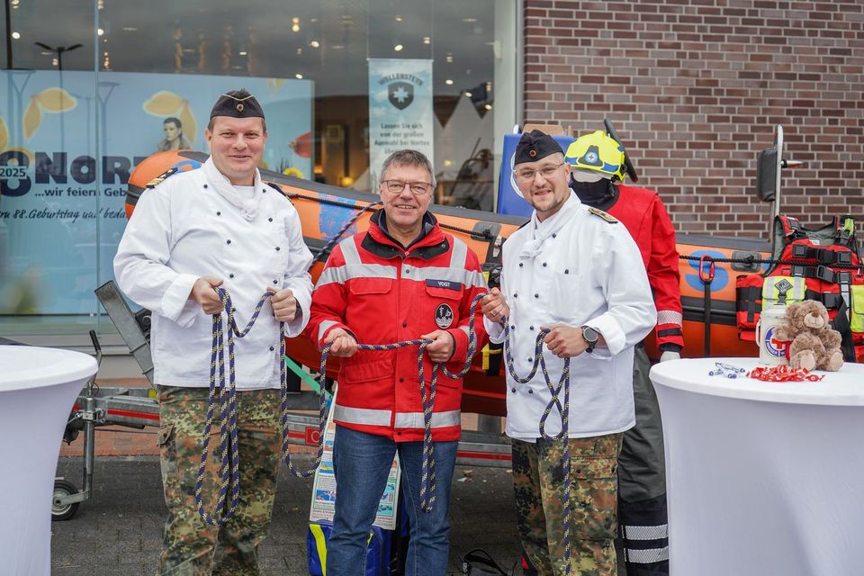 Drei Männer stehen vor einem Rettungsboot. Zwei von ihnen tragen eine weiße Koch-Jacke, der dritte eine rote Einsatzjacke.