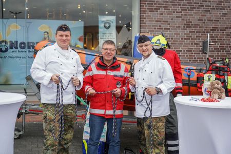 Drei Männer stehen vor einem Rettungsboot. Zwei von ihnen tragen eine weiße Koch-Jacke, der dritte eine rote Einsatzjacke.