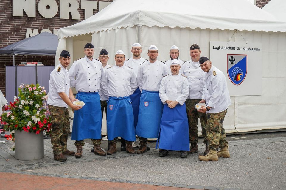 10 Marine-Köche in weißen Koch-Uniformen stehen vor einem weißen Zelt.