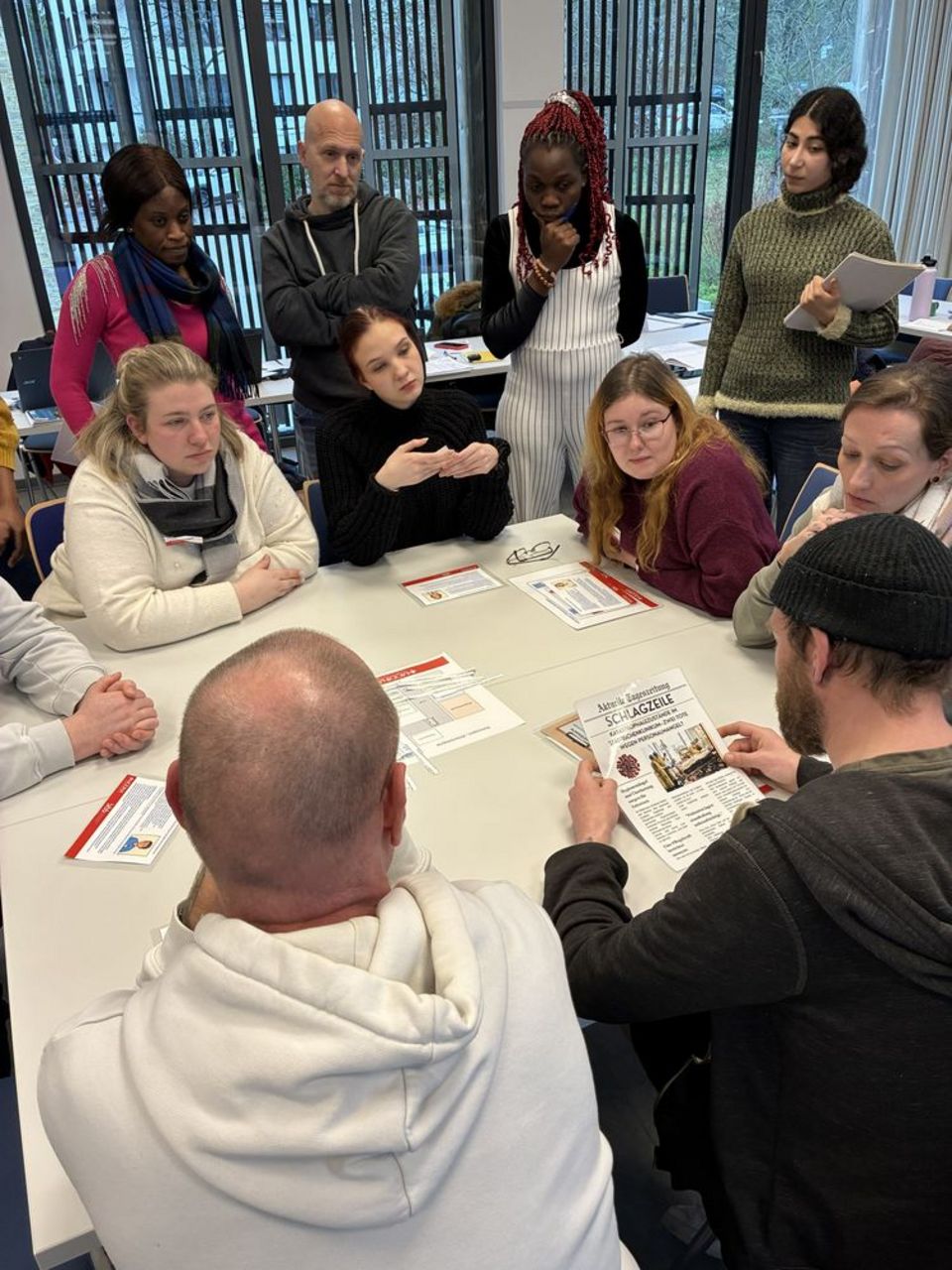 Zehn Personen sitzen und stehen um einen Tisch herum und besprechen sich.