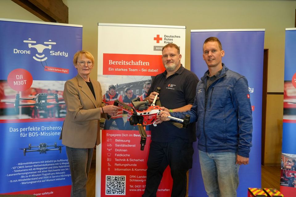 Drei Menschen posieren mit einer Drohne in den Händen vor Werbebannern. Eines zeigt das Logo des Deutschen Roten Kreuzes, das andere das Logo der Firma Drone Safety.