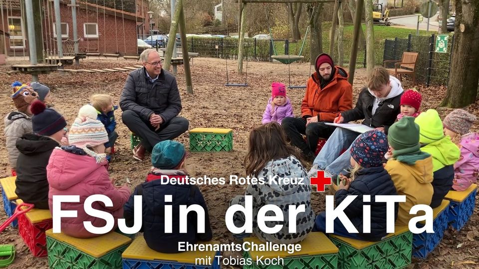 Auf einem Spielplatz sitzt eine Kita-Gruppe im Halbkreis. Dazwischen sitzen auch Erwachsene.