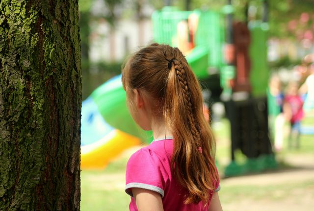 Blick von hinten auf ein kleines Mädchen, das neben einem Baum abseits eines Spielplatzes die anderen Kinder beobachtet.