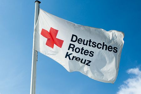 Im Wind weht eine weiße Flagge. Sie zeigt ein rotes Kreuz. Daneben steht in schwarzen Buchstaben: Deutsches Rotes Kreuz.