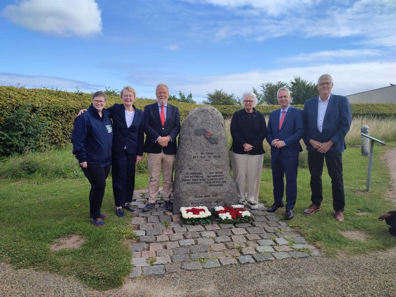 Je drei Personen stehen links und rechts eines Gedenksteins. Vor ihm liegen zwei Blumengestecke.