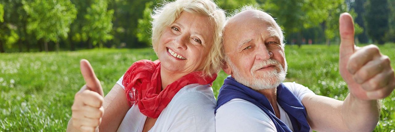Ein älteres Ehepaar sitzen auf einer Wiese und lehnt Rücken an Rücken. Beide lächeln und halten den Daumen hoch.