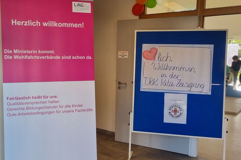 Links steht ein Roll-Up. Auf der oberen Hälfte steht weiß auf pink: Herzlich Willkommen! Die Ministerin kommt. Die Wohlfahrtsverbände sind schon da. Darunter steht pink auf weiß: Faislässlich heißt für uns: Qualitätsversprechen halten, Gerechte Bildungschancen für alle Kinder, Gute Arbeitsbedingungen für unsere Fachkräfte. Rechts neben dem Roll-Up zeigt ein Plakat: Herzlich Willkommen in der DRK-Kita Zeisigring.