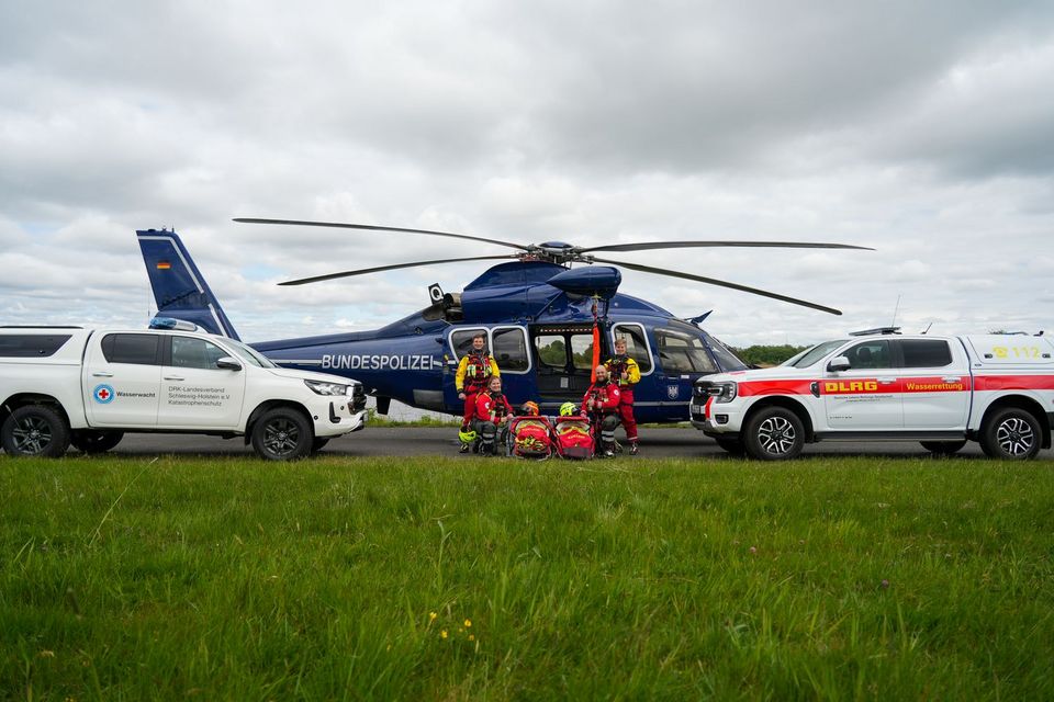 Vor einem blauen Bundespolizei-Hubschrauber stehen je zwei Einsatzkräfte von DRK und DLRG. Links der Gruppe steht ein weißer Pick-Up mit der Aufschrift "Wasserwacht". Rechts der Gruppe steht ein weiter mit der Aufschrift "DLRG".