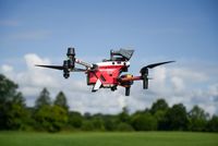 Eine rot-weiße DRK-Drohne im Flug. Im Hintergrund: wolkiger blauer Himmel sowie grüne Bäume am Rande einer Wiese