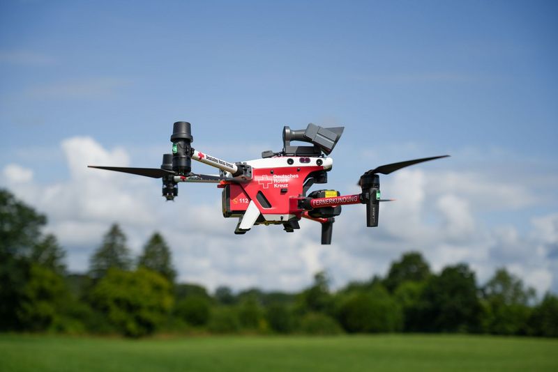Eine rot-weiße DRK-Drohne im Flug. Im Hintergrund: wolkiger blauer Himmel sowie grüne Bäume am Rande einer Wiese