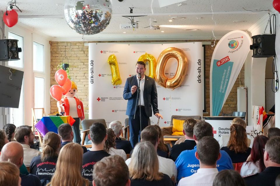 Ein junger Mann im Anzug steht auf einer bunt-geschmückten Bühne und spricht in ein Mikrofon. Vor der Bühne sitzen zahlreiche Menschen.