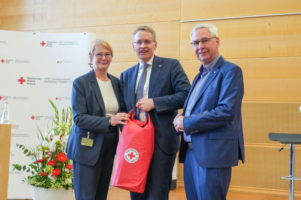 Drei Personen stehen nebeneinander auf einer Bühne und lächeln in die Kamera. Die mittlere Person hält einen roten DRK-Rucksack in der Hand.