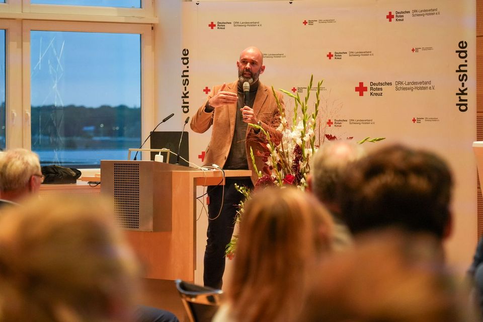 Clemens Beisel auf dem Parlamentarischen Abend des DRK SH Ein Mann im Anzug steht an einem Rednerpult und spricht zu einigen Menschen.