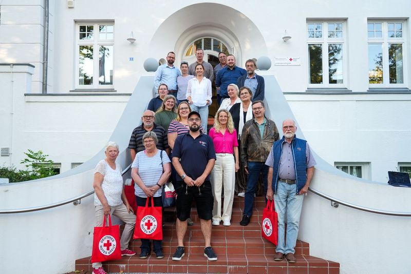Gruppenbild: 19 Personen stehen auf einer Treppe vor einem großen weißen Gebäude und schauen in die Kamera.