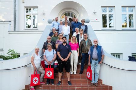 Gruppenbild: 19 Personen stehen auf einer Treppe vor einem großen weißen Gebäude und schauen in die Kamera.