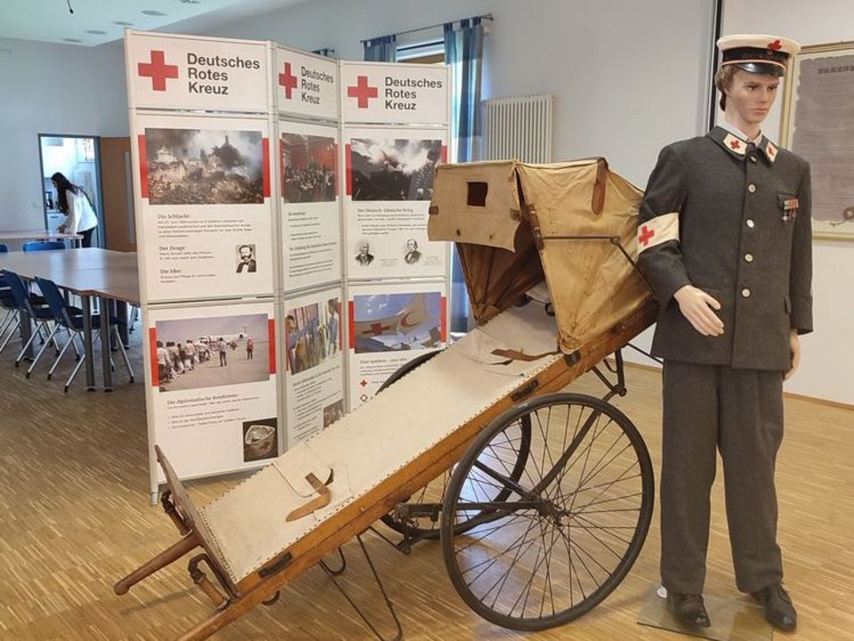 In einem Saal stehen große DRK-Stellwände mit Infos zur Geschichte des humanitären Völkerrechts. Davor steht eine alte hölzerne Patientenliege mit Rädern. Daneben trägt eine Puppe eine graue Uniform mit zahlreichen Rotkreuz-Symbolen.