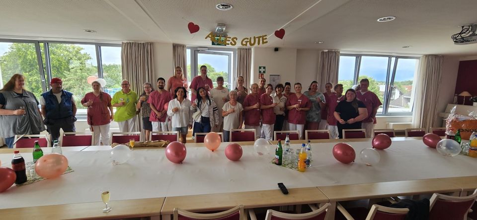 Gut 20 Menschen stehen in einem geschmückten Saal. Auf den Tischen sind Ballons und an der Decke hängt eine "Alles Gute"-Girlande.