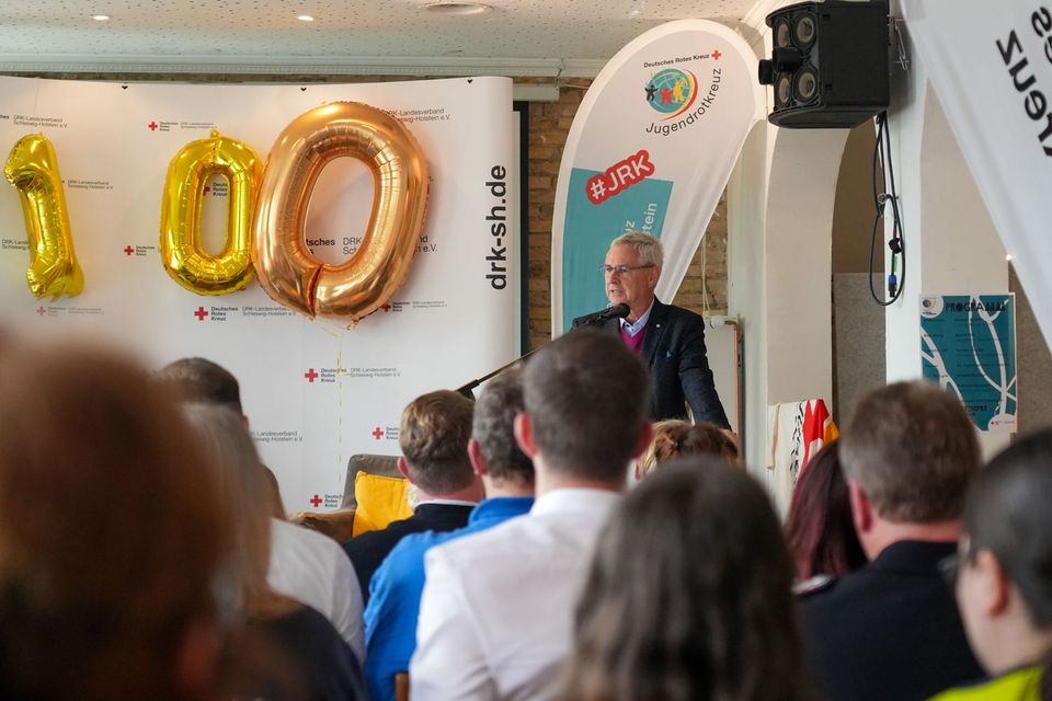 Ein älterer Mann in Anzug steht an einem Rednerpult und spricht zu zahlreichen Menschen.