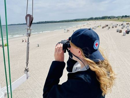 Eine Frau steht auf einem Wachturm und guckt durch ein Fernglas auf den Strand. Auf dem Strand sind Menschen und Meer in der Ferne zu erkennen.