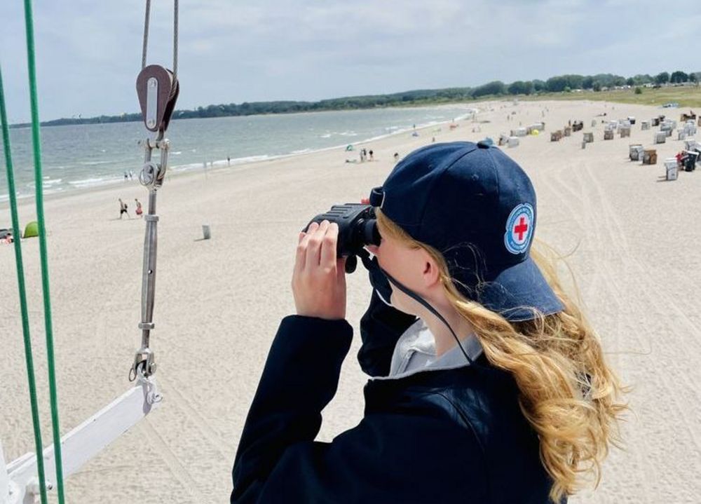 Eine Frau steht auf einem Wachturm und guckt durch ein Fernglas auf den Strand. Auf dem Strand sind Menschen und Meer in der Ferne zu erkennen.