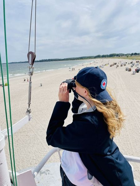 Eine Frau steht auf einem Wachturm und guckt durch ein Fernglas auf den Strand. Auf dem Strand sind Menschen und Meer in der Ferne zu erkennen.
