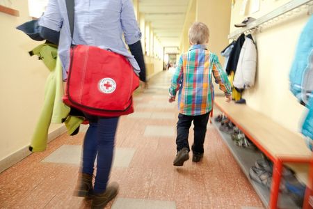 Blick von hinten auf ein kleines Kind, das einen Schulflur entlang geht. Schräg links dahinter geht eine erwachsene Person, die eine rote Tasche mit DRK-Logo trägt.