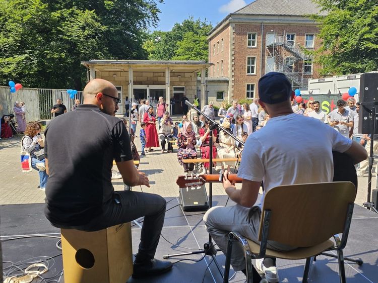 Blick von hinten auf zwei Personen, die auf einer Bühne Cajon und Gitarre spielen. Im Hintergrund erkennt man das Publikum: Rund 50 Leute.