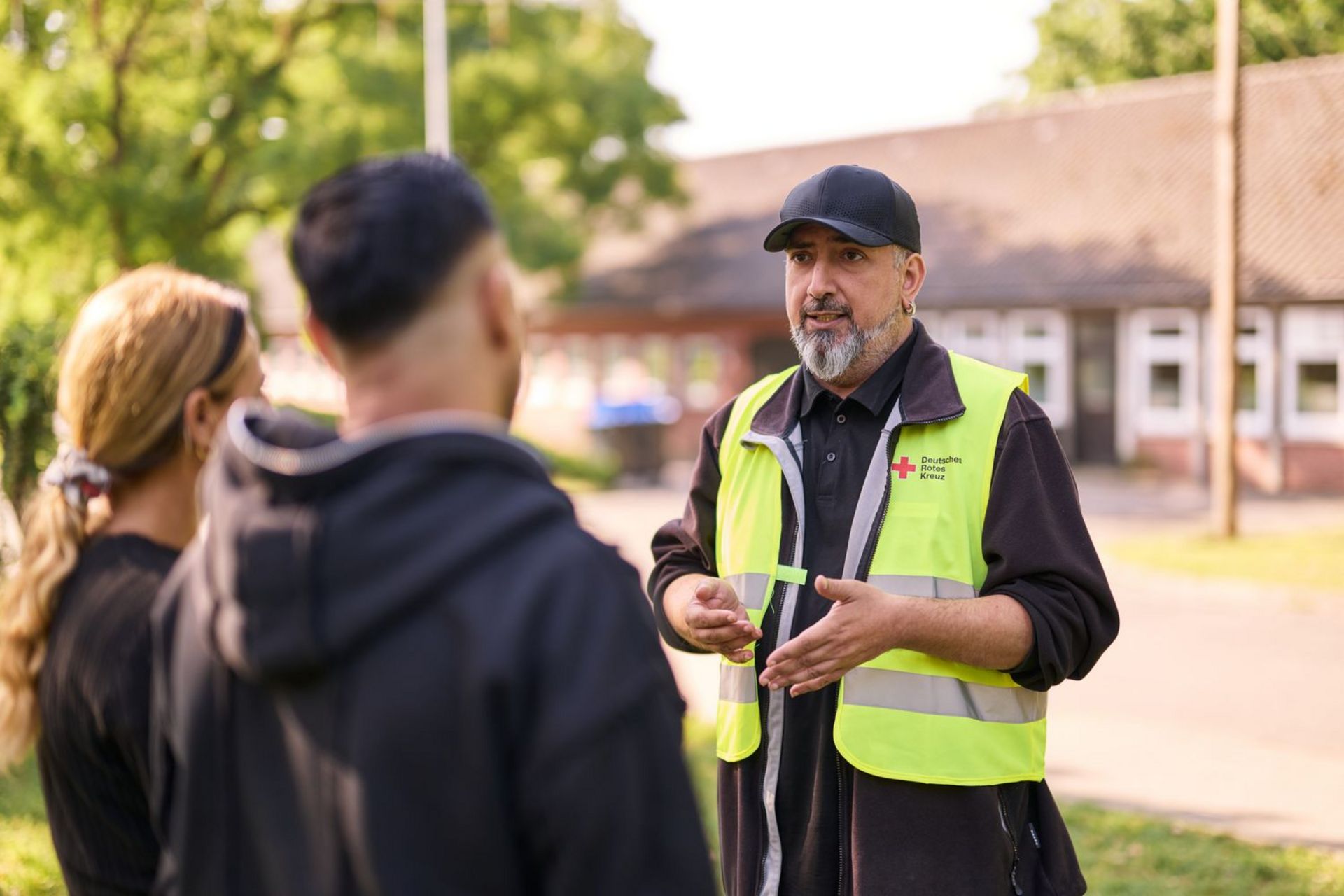 Drei Personen unterhalten sich. Eine von ihnen trägt eine gelbe Warnweste mit DRK-Logo.