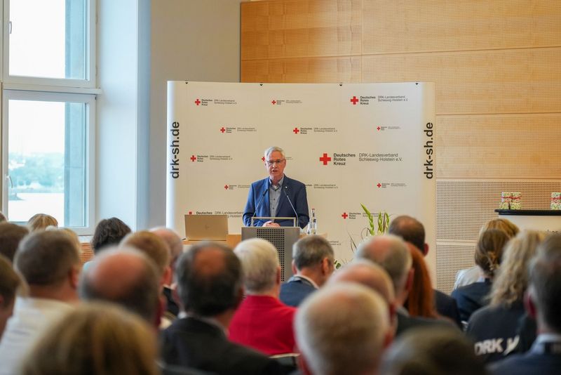 Ein Mann steht an einem Rednerpult und spricht zu großem Publikum im Vordergrund.