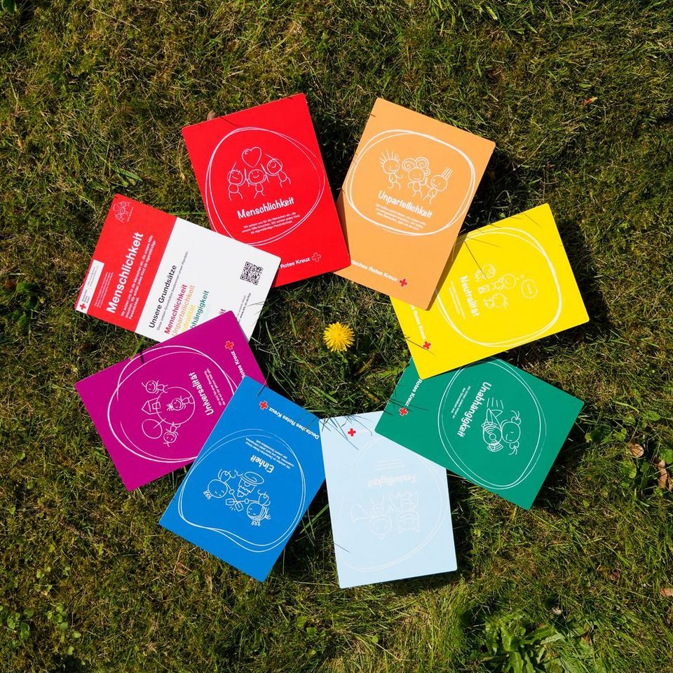 Auf grünem Gras liegen 8 bunte Din A 5 Karten um eine gelbe Löwenzahnblüte herum. Auf den Karten stehen die Rotkreuz-Grundsätze.