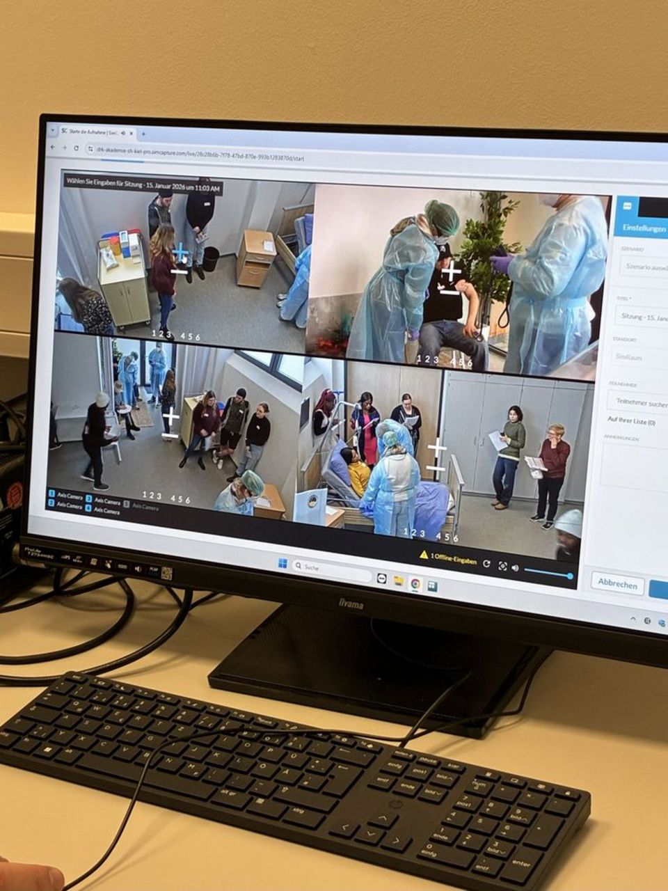 Ein Bildschirm zeigt vier Blickwinkel von Kameras in einem Raum, in dem Pflegekräfte in Schutzanzügen eine Patientin betreuen.