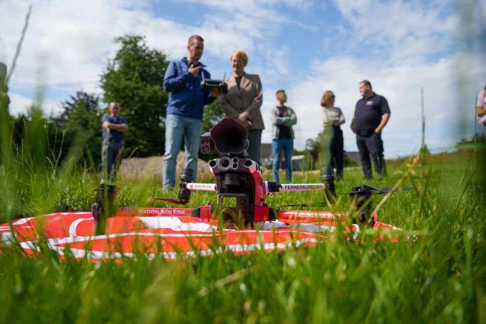 Froschperspektive auf einer Wiese: Eine weiß-rote Drohne mit DRK-Logos steht auf dem Boden. Im Hintergrund stehen sechs Menschen. Zwei von ihnen schauen auf eine Fernbedienung.